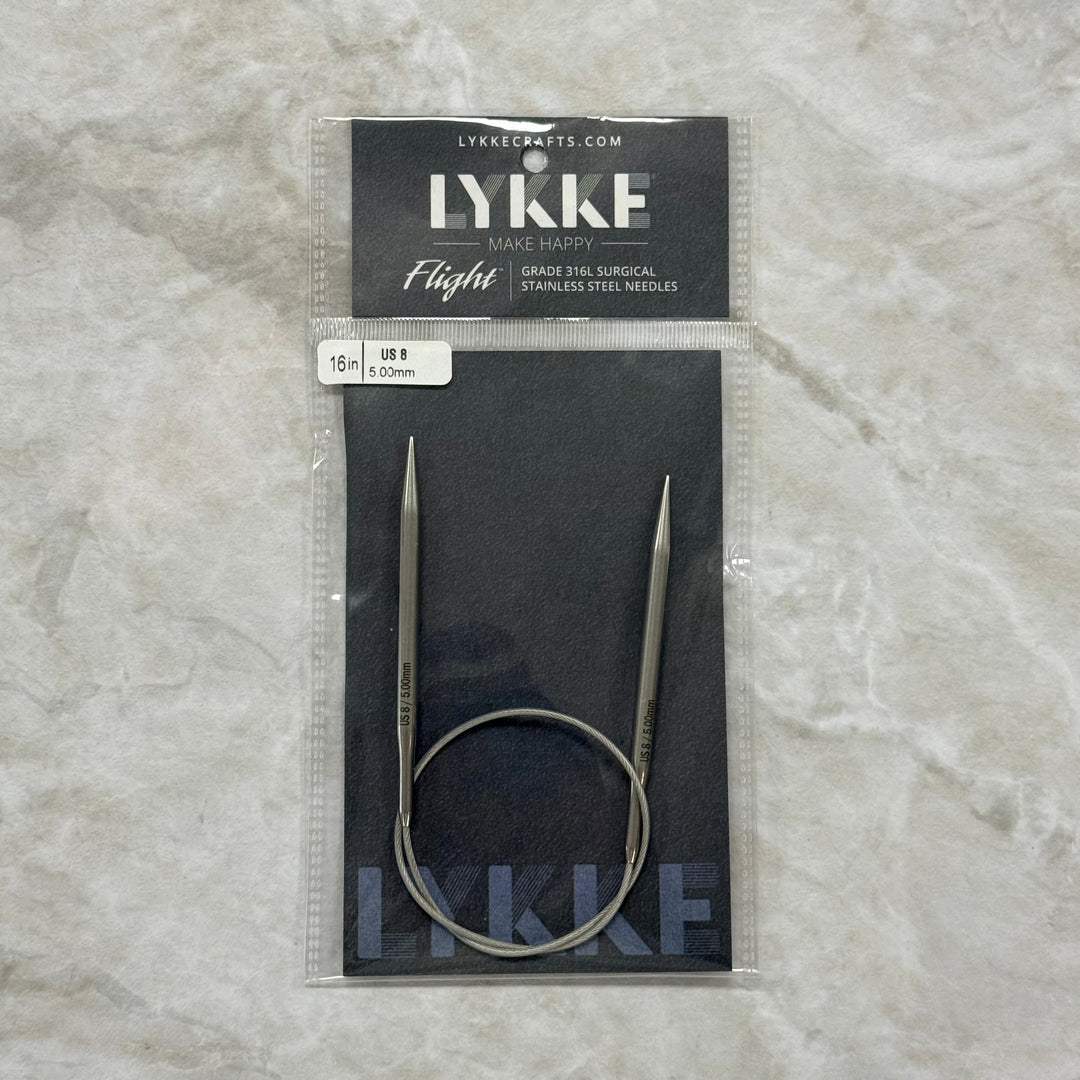 Lykke Flight circular knitting needles in their original packaging, displayed on a textured light gray surface. The needles are made of stainless steel and feature a continuous loop design.