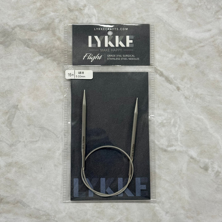 Lykke Flight circular knitting needles in their original packaging, displayed on a textured light gray surface. The needles are made of stainless steel and feature a continuous loop design.