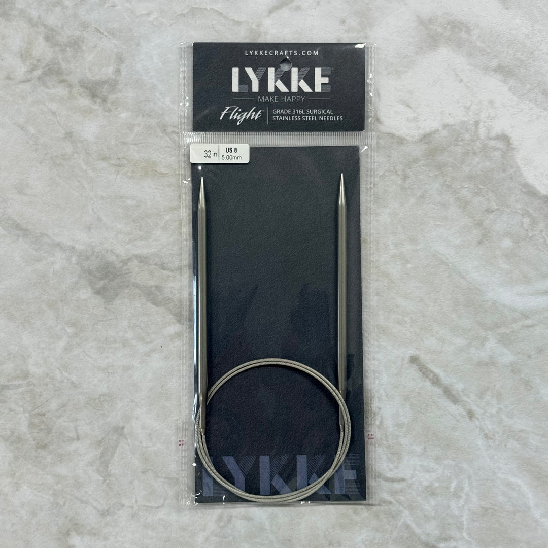 Lykke Flight circular knitting needles in their original packaging, displayed on a light gray marble surface. The needles are made of stainless steel and come with a black circular cable and a black display card.