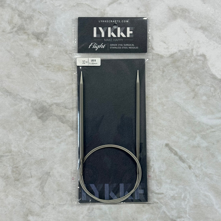 Lykke Flight circular knitting needles in their original packaging, displayed on a light gray marble surface. The needles are made of stainless steel and come with a black circular cable and a black display card.